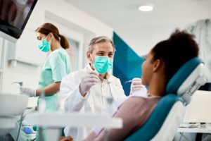 Woman with brown hair in teal dentist chair while dentist in white coat and green mask holds up instruments and an aide prepares tools behind him